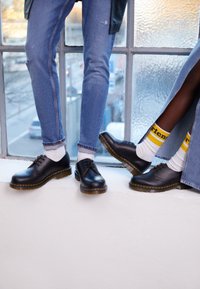 Pantofi din piele neagră cu cusături galbene, asortați cu șosete albe și blugi. Unul dintre pantofi are o finisare netedă; celălalt are o talpă texturată.