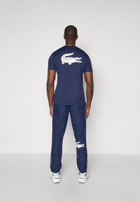 Camicia blu navy a maniche corte con un grande logo bianco di coccodrillo sul retro, abbinata a pantaloni sportivi blu navy e sneakers bianche.