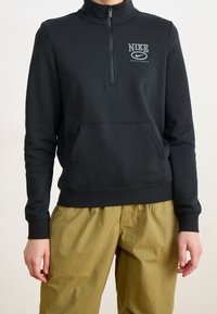 Svart Nike huvtröja med hög krage, halvdagsdragkedja, framficka i kängurustil och vitt logotyp på bröstet. Mjuk tygtextur.