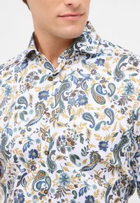 Witte blouse met een paisley- en bloemenpatroon in blauwe, groene en gouden tinten; heeft een knoopsluiting en een puntige kraag.
