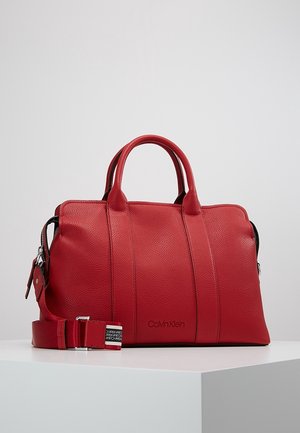Rote Lederhandtasche von Calvin Klein mit doppelten Griffen und abnehmbarem Schulterriemen auf weißer Oberfläche vor grauem Hintergrund.