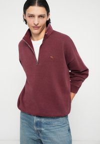 Pull en polaire bourgogne avec col zippé mi-haut et logo brodé, associé à un jean en denim bleu clair. Texture douce et coupe décontractée.