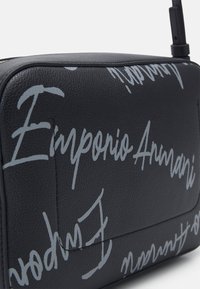 Black textured leather bag with gray cursive "Emporio Armani" script printed across its surface and a zipper at the top.