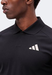 Gros plan d'une personne portant une chemise noire à col avec un logo blanc à trois barres sur la poitrine gauche sur fond uni.