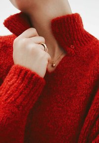 Suéter rojo de punto con un alto cuello acanalado y detalle de botones. Mano que lleva un anillo dorado y un collar con un colgante en forma de corazón.