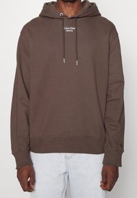 Sudadera marrón hecha de tela suave, con un bolsillo frontal, capucha con cordones y un logo bordado en el pecho y la manga.