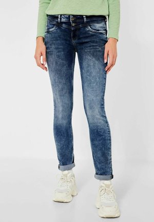 Jeans ajustados de color azul con un lavado desvaído, cierre de dos botones, bolsillos frontales y puños enrollados, combinados con zapatillas blancas.