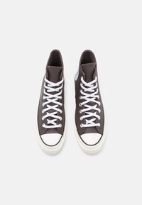 Brown high-top sneakers with white laces, rubber toe cap, and a circular logo patch on the side. Smooth fabric texture, classic design.