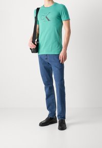 Grön bomullströja med en mörk 'CK'-logotyp, parat med blå denimjeans och svarta läderloafers, samt en svart axelremsväska.