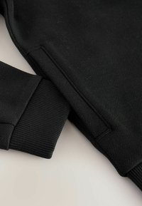 Sudadera negra con tejido texturizado, puños acanalados y costuras. El diseño tiene un aspecto simple y limpio, sin logotipos ni adornos visibles.