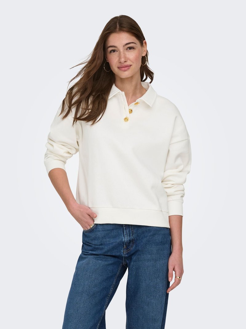 Jeune femme aux longs cheveux bruns portant des boucles d'oreilles anneaux dorées, un polo à manches longues blanc avec des boutons dorés et un jean bleu, debout avec une main dans la poche.