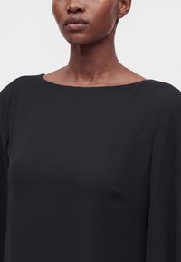 Femme portant un haut noir à encolure bateau, montrant son cou, ses épaules et le haut de sa poitrine sur un fond uni gris clair.