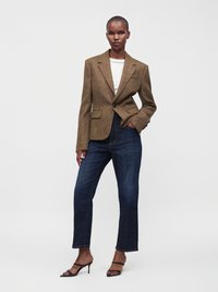 Femme portant un blazer marron à un bouton, un haut blanc, un jean bleu foncé et des sandales à talons noires, debout avec une main dans la poche.