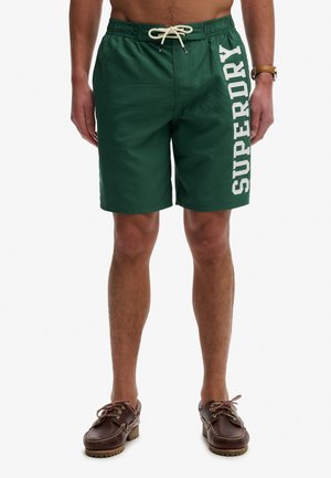 Hombre vistiendo pantalones cortos de baño verde oscuro con texto blanco "SUPERDRY" en una pierna y zapatos náuticos de cuero marrón, de pie contra un fondo blanco.