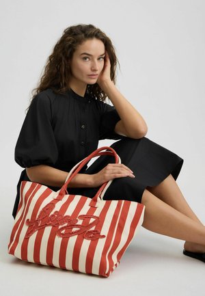 Borsa a tracolla in tela a righe rosse e bianche con ricami texturizzati. Presenta manici robusti e un design spazioso, appoggiata accanto a un modello seduto.