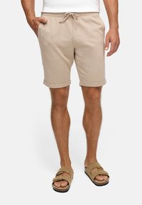 Shorts beiges à texture avec une taille à cordon de serrage et des ourlets retournés, associés à des sandales marron à lanières sur des pieds nus.