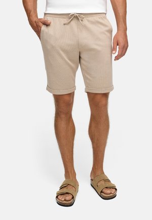 Pantalones cortos beige, texturizados, con cintura de cordón y puños enrollados, combinados con sandalias marrones de tiras sobre pies descalzos.