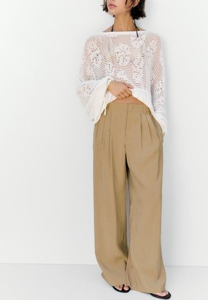 Donna con pantaloni beige a gamba larga e top bianco in crochet a maniche lunghe, in piedi davanti a uno sfondo bianco uniforme.