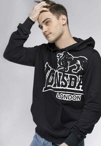 Sudadera negra con bolsillo canguro, presenta un diseño gráfico blanco que dice "LONSDALE LONDON" en el frente, y tela texturizada.