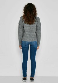 Femme aux cheveux bouclés portant un pull gris en tricot, un jean skinny bleu et des baskets noires, debout face à un mur blanc uni.