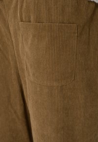 Καφέ υφαντό κοτλέ με κάθετες ραβδώσεις, που διαθέτει τσεπάκι με πατς στο πίσω μέρος. Μαλακή υφή και ζεστό, γήινο χρώμα.