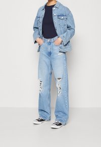 Person wearing light blue ripped wide-leg jeans, matching denim jacket, black shirt, and black-and-white sneakers, standing with hands in pockets.