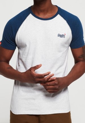 Mann trägt ein weißes und dunkelblaues Raglan-T-Shirt mit kleinem Logo auf der linken Brust, hält die Hände vor dem Oberkörper verschränkt.