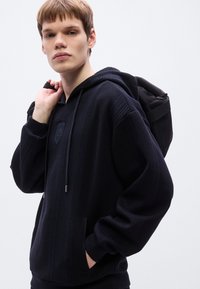 Μαύρο φούτερ με ανάγλυφη υφή, ριμπ μοτίβο, κορδόνι και τσέπη καγκουρό. Μοντέλο που φοράει μαύρο σακίδιο πλάτης, ουδέτερο φόντο.