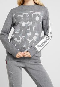 Felpa grigia con grande stampa "SuperDry" in bianco e camouflage grigio. Maniche lunghe con polsini; pantaloni grigi abbinati visibili.