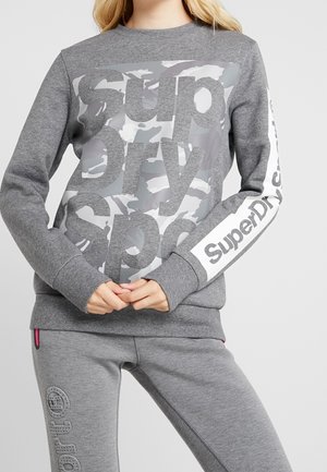 Felpa grigia con grande stampa "SuperDry" in bianco e camouflage grigio. Maniche lunghe con polsini; pantaloni grigi abbinati visibili.