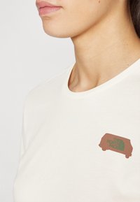 Krémové bavlněné tričko s kulatým výstřihem, s hnědým a zeleným logem The North Face umístěným na levé části hrudi.