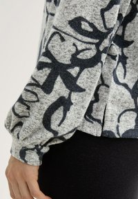 Grauer Pullover mit dunkelbenten Blumenmuster, weicher Textur und gerafften Ärmeln. Mit seitlichen Schlitzen am Saum. Darunter schwarze Leggings.