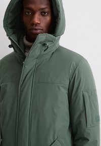 Zwarte man in een groene winterjas met capuchon en rits aan de voorkant, met een hoge kraag, staand tegen een eenvoudige lichte muur.