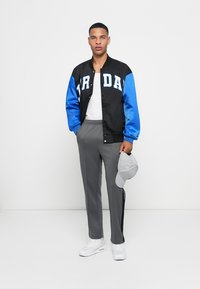 Sort og blå varsityjakke med hvidt tryk, grå sportsbukser, hvide sneakers og en grå kasket, stående foran en hvid baggrund.
