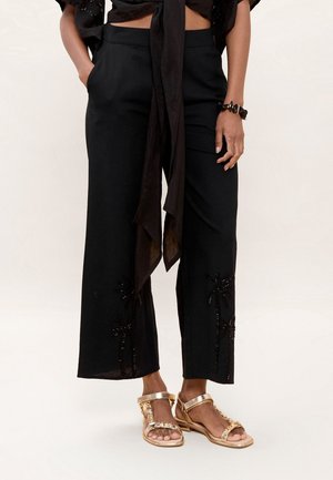 Mujer con pantalones negros de pierna ancha con un sutil bordado de palmeras y sandalias planas doradas, de pie con las manos en los bolsillos.