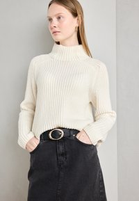 Pulover crem cu guler înalt și nervuri, cu mâneci lungi, asortat cu o fustă din denim negru și o curea neagră cu o cataramă circulară aurie.