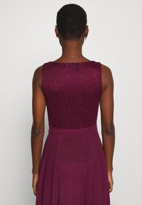 Femme aux cheveux courts portant une robe sans manches en dentelle violet foncé, vue de dos sur un fond clair uni.