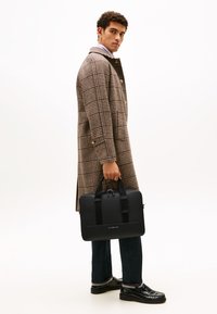 Black laptop bag with simple hardware, carried by a person in a checked brown coat, dark jeans, and shiny black shoes.