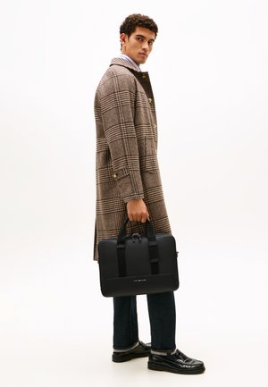 Black laptop bag with simple hardware, carried by a person in a checked brown coat, dark jeans, and shiny black shoes.