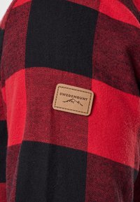Tissu à carreaux rouge et noir avec un patch rectangulaire beige étiqueté « Swedemount » et un logo de montagne brodé dessus.