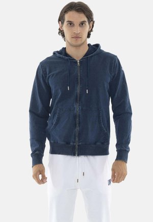Man draagt een marineblauwe zip-up hoodie met voorzakken en witte broek met trekkoord, staand gezicht naar voren.