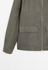 Brun tekstureret stofjakke med lynlås foran og to store klaplommer tæt på kanten, der viser venstre ærme og side.