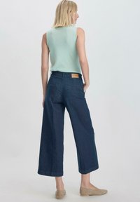 Donkere denim broek met wijde pijpen en een bruin leren patch aan de achterkant, gecombineerd met een lichtblauwe mouwloze top. Glad stoffen textuur.