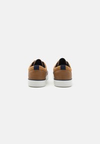 Brown suede sneakers with a smooth finish, white rubber sole, and navy accents at the heel and tongue. Back view shows slip-on design.