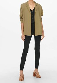 Donna che indossa un blazer beige, una maglietta nera con bottoni, pantaloni skinny neri e sandali con tacco blocco e fasce beige su uno sfondo bianco.