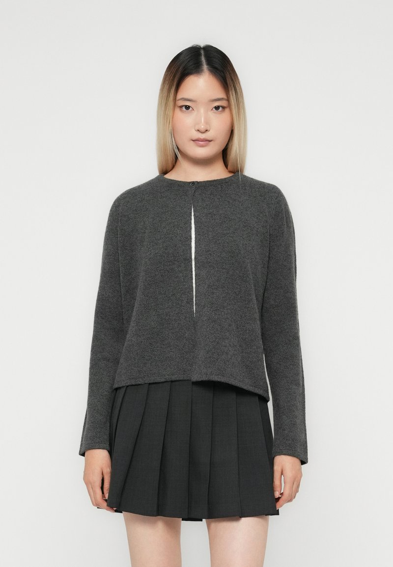 Jeune femme avec des cheveux raides dégradés, portant un cardigan boutonnière gris foncé sur un haut blanc et une jupe plissée noire, se tenant devant un fond blanc.