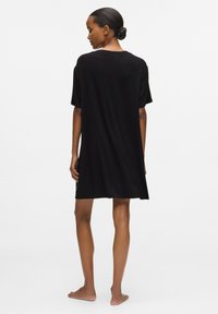 Femeie stând desculță, cu fața spre spate, purtând o rochie neagră lejeră cu mâneci scurte, părul prins într-un coc jos, pe fundal alb.