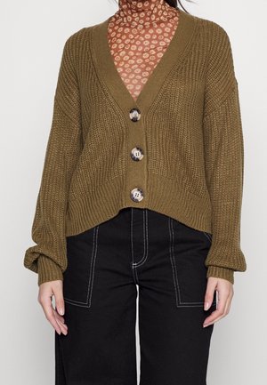 Person iført en olivengrøn strikket cardigan med store knapper over en brun blomstret højhalset trøje og sorte bukser med hvide syninger.