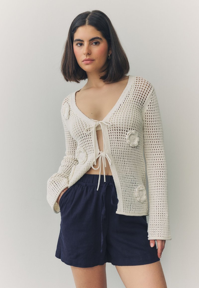 Gebreid vest in roomwit met een V-hals, striksluiting en bloemapplicaties; gecombineerd met marineblauwe shorts. Opengeweven textuur.