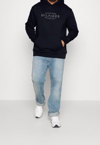 Felpa navy con tasca frontale e marchio "Hilfiger", abbinata a jeans azzurri e sneakers bianche, con maniche arrotolate.